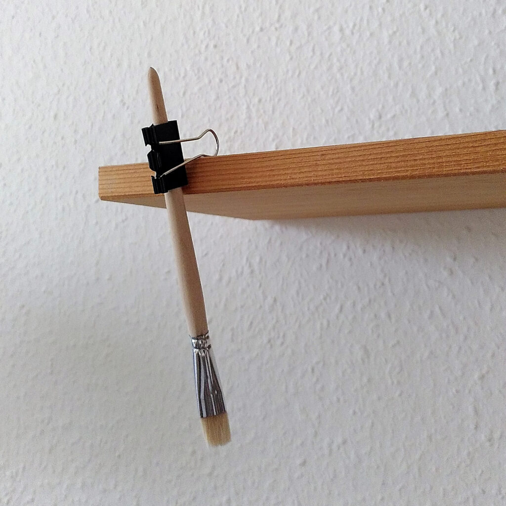 Pinsel, dessen Stiel in eine Foldback-Klammer eingeklemmt ist, die mit ihren Bügel am Rand eines Regalbretts hängt.