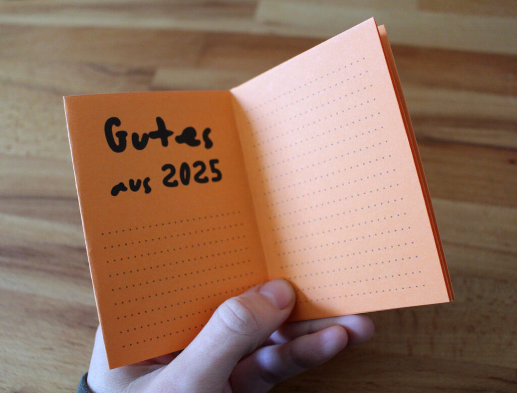 Eine Hand hält ein aufgeschlagenes Zine aus orangefarbenem Papier, auf der Innenseite kann man die Überschrift "Gutes aus 2025" lesen